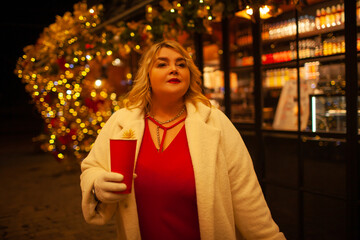 Plus size woman walks around the city during the Christmas of the week with a hot drink in a glass against the background of flickering light bulbs