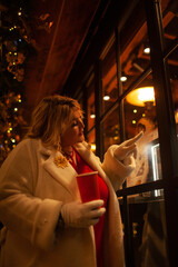 Plus size woman walks around the city during the Christmas of the week with a hot drink in a glass against the background of flickering light bulbs