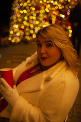 Plus size woman walks around the city during the Christmas of the week with a hot drink in a glass against the background of flickering light bulbs