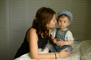 Loving mother admiring her baby boy dressed in elegant outfit and cap, sitting on a changing table...