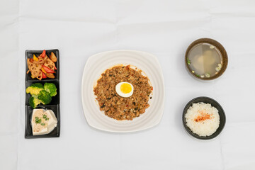 A Thai basil minced pork set meal arranged on a tray, including a portion of stir-fried spicy basil pork, a bowl of steamed white rice, three side vegetables, and a bowl of soup. The presentation refl