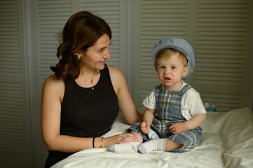 Loving mother admiring her baby boy dressed in elegant outfit and cap, sitting on a changing table...