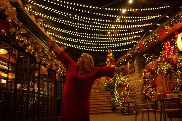Plus size woman walks around the city during the Christmas of the week with a hot drink in a glass against the background of flickering light bulbs