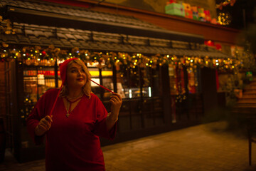 Plus size woman walks around the city during the Christmas of the week with a hot drink in a glass against the background of flickering light bulbs