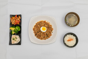 A Thai basil minced pork set meal arranged on a tray, including a portion of stir-fried spicy basil pork, a bowl of steamed white rice, three side vegetables, and a bowl of soup. The presentation refl