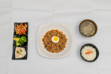 A Thai basil minced pork set meal arranged on a tray, including a portion of stir-fried spicy basil pork, a bowl of steamed white rice, three side vegetables, and a bowl of soup. The presentation refl