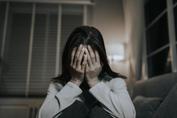 A young woman sits alone on a sofa at home, looking sad and contemplative, reflecting emotional...