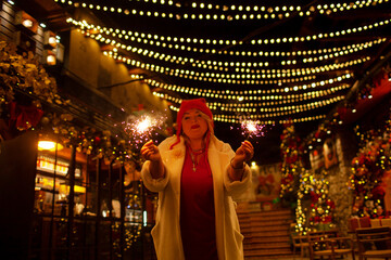 Plus size woman walks around the city during the Christmas of the week with a hot drink in a glass against the background of flickering light bulbs