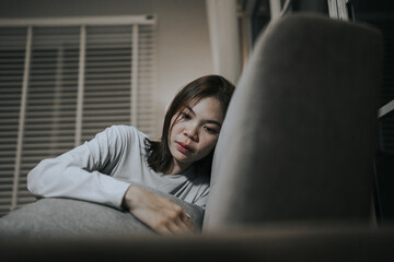 A young woman sits alone on a sofa at home, looking sad and contemplative, reflecting emotional...