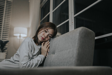 A young woman sits alone on a sofa at home, looking sad and contemplative, reflecting emotional...