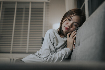 A young woman sits alone on a sofa at home, looking sad and contemplative, reflecting emotional...