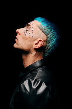 Side view of serene male model with eye stickers on face and dyed blue hair on black background in studio