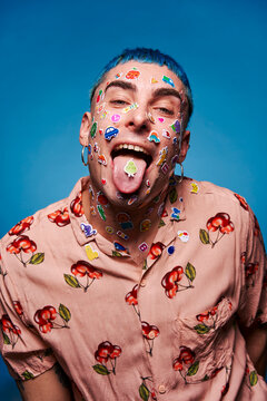 Eccentric male model with blue hair and stickers on face looking at camera and showing tongue in studio