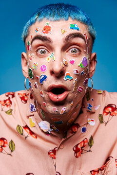 Eccentric male model with blue hair and stickers on face looking at camera and showing tongue in studio