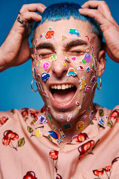 Screaming male model with blue hair and stickers on face looking at camera and showing tongue in studio