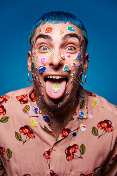Eccentric male model with blue hair and stickers on face looking at camera and showing tongue in studio