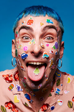 Naklejki Eccentric male model with blue hair and stickers on face looking at camera and showing tongue in studio