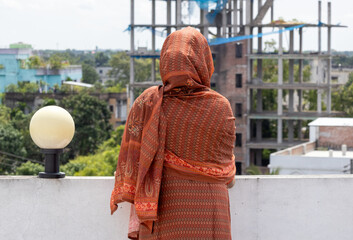 A Bengali Muslim woman in a modest brown salwar kameez with her head veiled, standing alone on a rooftop. Her back is to the viewer as she quietly observes the morning outdoor scene.
