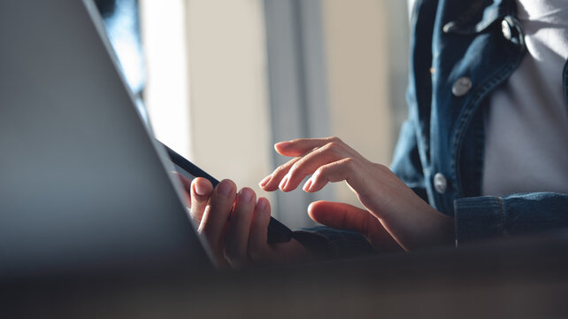 Closeup, business woman sitting at table using mobile smrt phone during working on laptop computer at home office. Young asian woman freelancer  surfing the internet, online working from home
