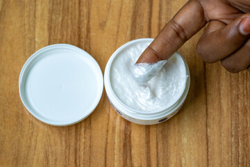 Woman's hand gently taking cream from an opened cosmetic jar. The lid rests nearby on a rustic wooden surface. Ideal for beauty, wellness, and self-care content.