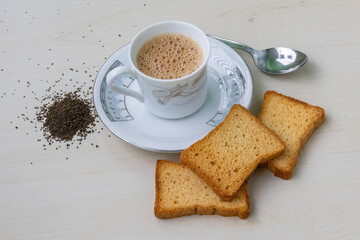 Hot, frothy milk tea in a teacup with golden rusk biscuits. Fresh and dried tea leaves scattered on...