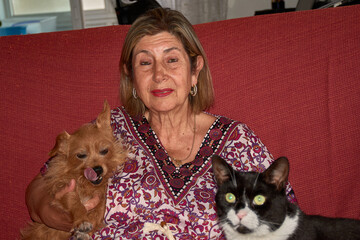 An elderly woman shares a sofa with her small ginger dog and her large black cat in a warm, homey atmosphere.