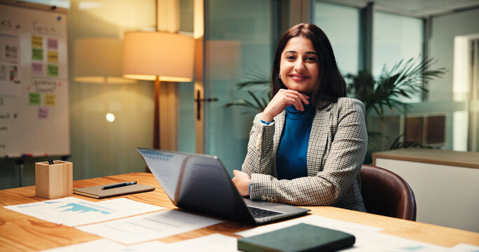 Laptop, portrait and statistics with business woman in office at night for social media analytics. Computer, development and smile of happy Indian employee at desk in workplace for evening deadline - Powered by Adobe