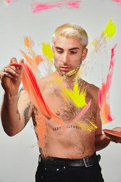 Stylish shirtless male model with palette smearing colorful paint on transparent glass against gray background