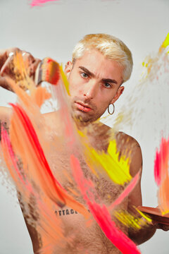 Stylish shirtless male model with palette smearing colorful paint on transparent glass against gray background