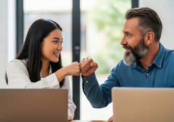 A young Asian woman and a mature man with a beard celebrate a success at work with a fist bump.