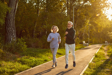 Healthy lifestyle. Happy senior husband and wife in sportive outfits running outdoors in city park. Active couple of retired people in sports clothes are doing regular jogging together in morning.