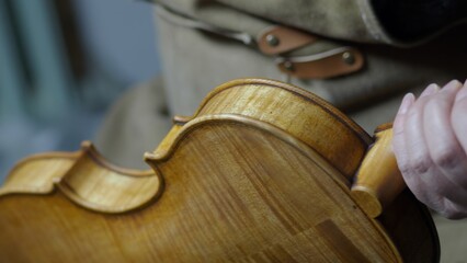 Craftsman's skilled hands applying amber toned varnish, meticulously antiquing violin body with...