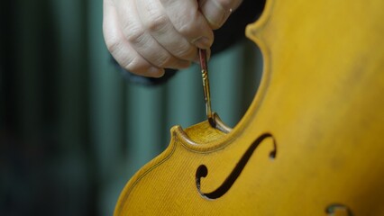 Experienced craftsman carefully brushing varnish onto violin, applying traditional antiquing method...