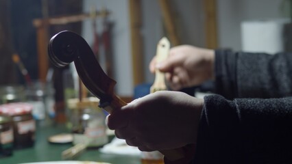 Skilled luthier carefully applying varnish to violin scroll, using brush and fingertip in workshop surrounded by woodworking tools and lutherie equipment