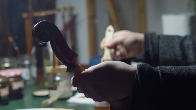Skilled luthier carefully applying varnish to violin scroll, using brush and fingertip in workshop surrounded by woodworking tools and lutherie equipment