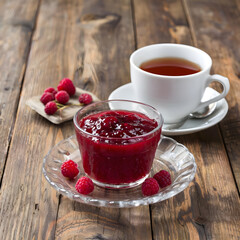 Raspberry jam in a glass and with tea in a white porcelain cup with fresh berries around on a wooden table. Light, cozy atmosphere of the morning and tea drinking.