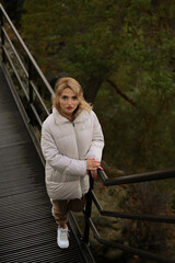 Young woman in a white puffer jacket walking through a beautiful green natural park on a sunny day. Outdoor lifestyle, peaceful moment, nature and fashion concept.