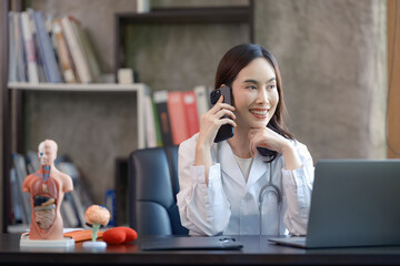 An Asian female doctor is talking on the phone with her patient while working in her hospital office.