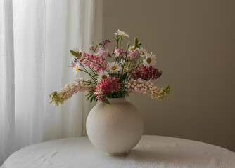 Bouquet of garden flowers of daisies, lupines in a round ceramic vase on a table with a white...
