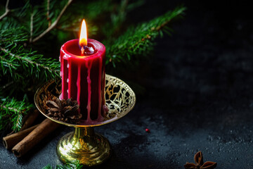 Close-up of an elegant red candle in vintage brass candleholder, melted wax dripping down the sides, evergreen sprigs and cinnamon sticks scattered around, dark moody background