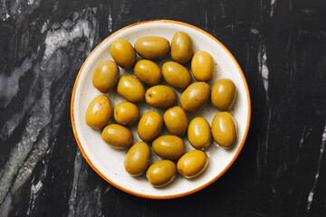 Bowl of green olives on dark marble background. Top view composition. Tasty Mediterranean snack. Healthy vegetarian food. Organic ripe olives in ceramic bowl. Simple rustic natural appetizer idea.