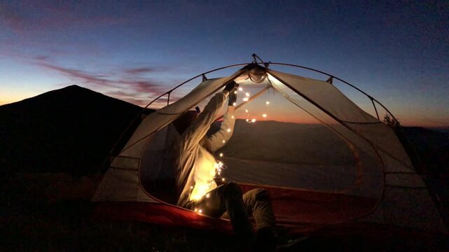 Woman untangling string lights during mountain camping at dusk inside a tent