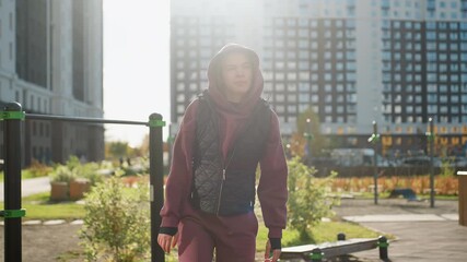 Gym goer in hoodie and vest stretches arms and legs outdoors in urban park with pedestrians passing by playground bars under morning sunlight preparing workout routine with focus and energy