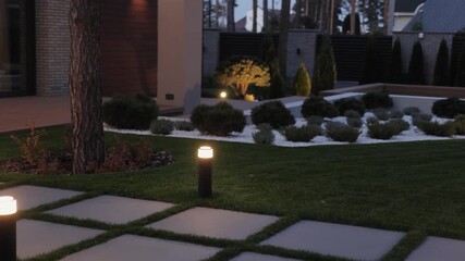 Landscaped garden at night. Lanterns illuminate the path with soft light. Street lamps in the floor on the grass. The warm glow highlights the lush greenery, creating a calming atmosphere.