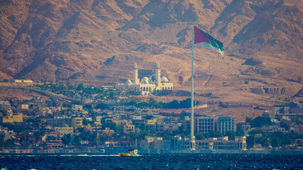 View of Aqaba city with Jordanian flag and mosque against desert mountains
