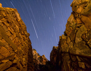 Star trails streak across the night sky above rugged rock walls in a narrow canyon, capturing the motion of the stars and the raw beauty of desert terrain.

