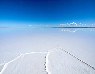 Vast salt flat under a clear blue sky with distant clouds mirrored on the surface. Cracked salt patterns in the foreground create a surreal, dreamlike landscape.