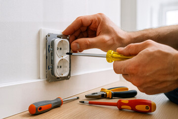 Close-up of electrician hands fixing a wall socket using a screwdriver, with tools on the floor in a home setting.