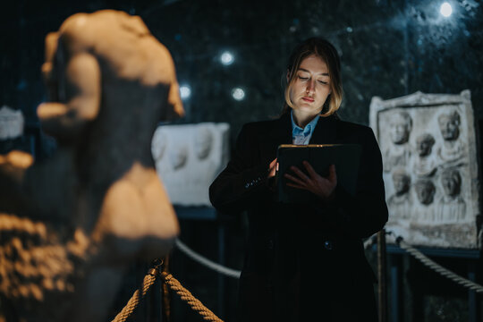 A woman in formal attire examines artifacts in a dimly lit museum, utilizing a digital device to document or research, creating an atmosphere of focus on history and cultural preservation.
