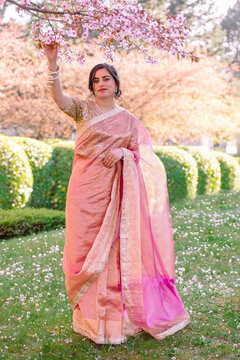 A woman in a shimmering pink traditional outfit stands serenely among cherry blossoms, radiating warmth, elegance, and cultural pride.